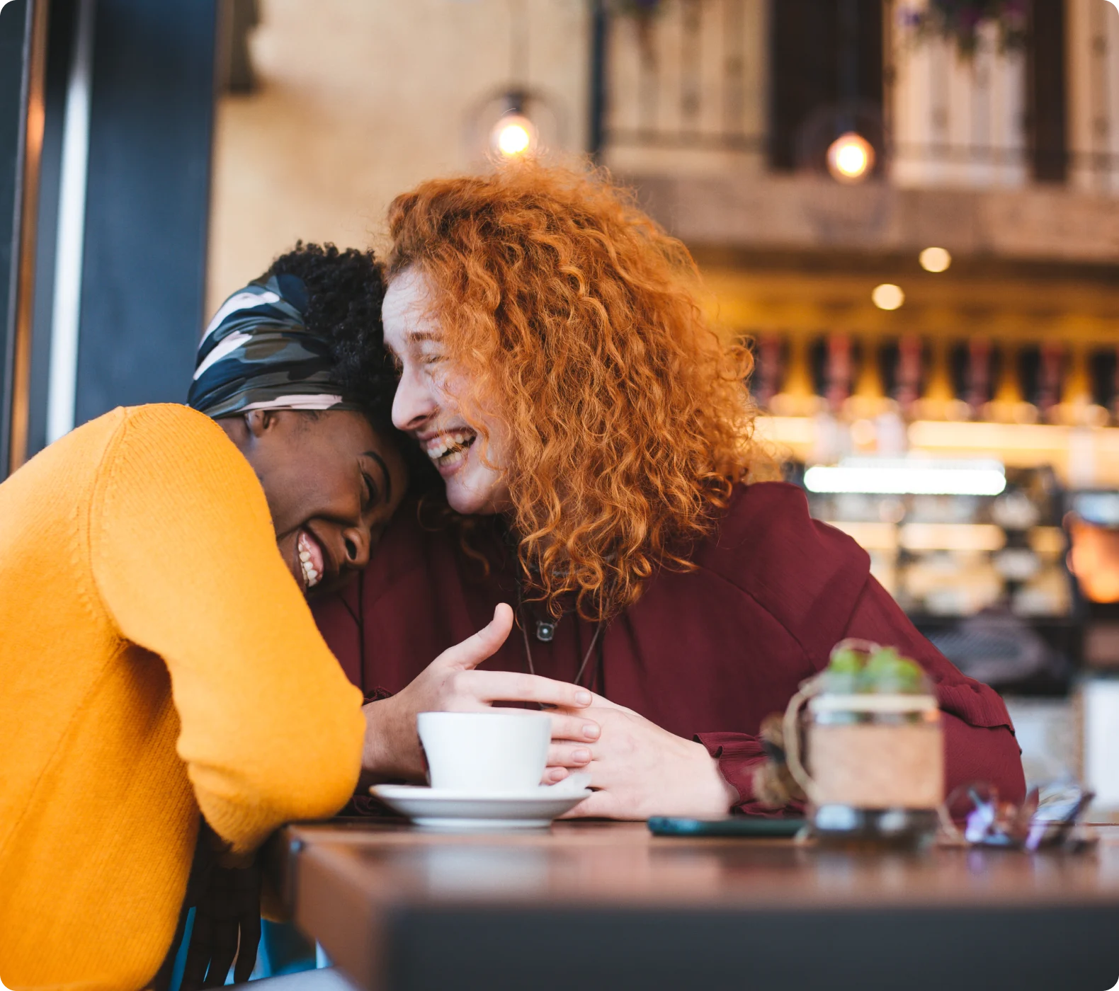 Two women at a coffee shop, their heads together, smiling