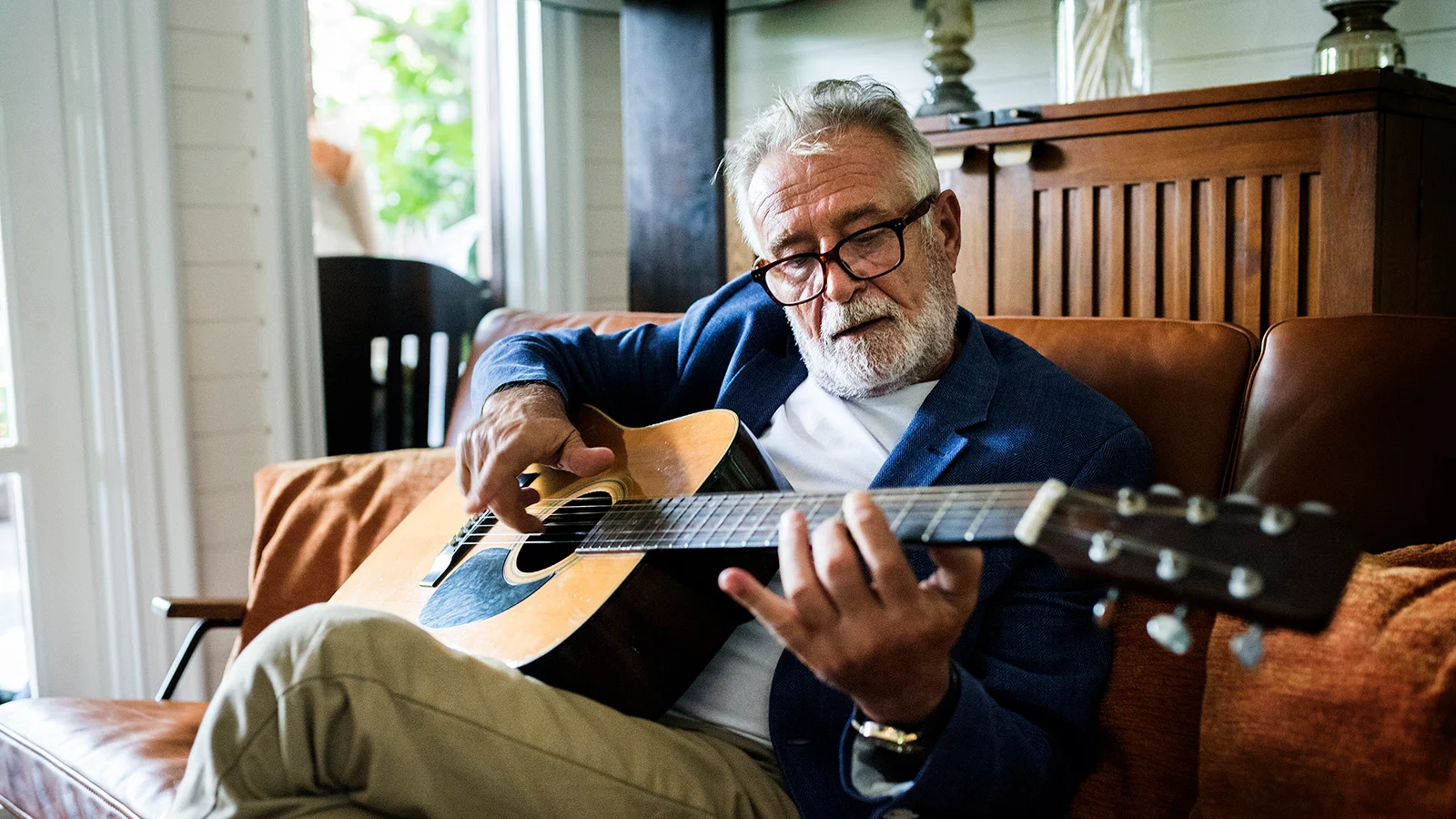 Person seated on a sofa playing an acoustic guitar in a sunlit living room.