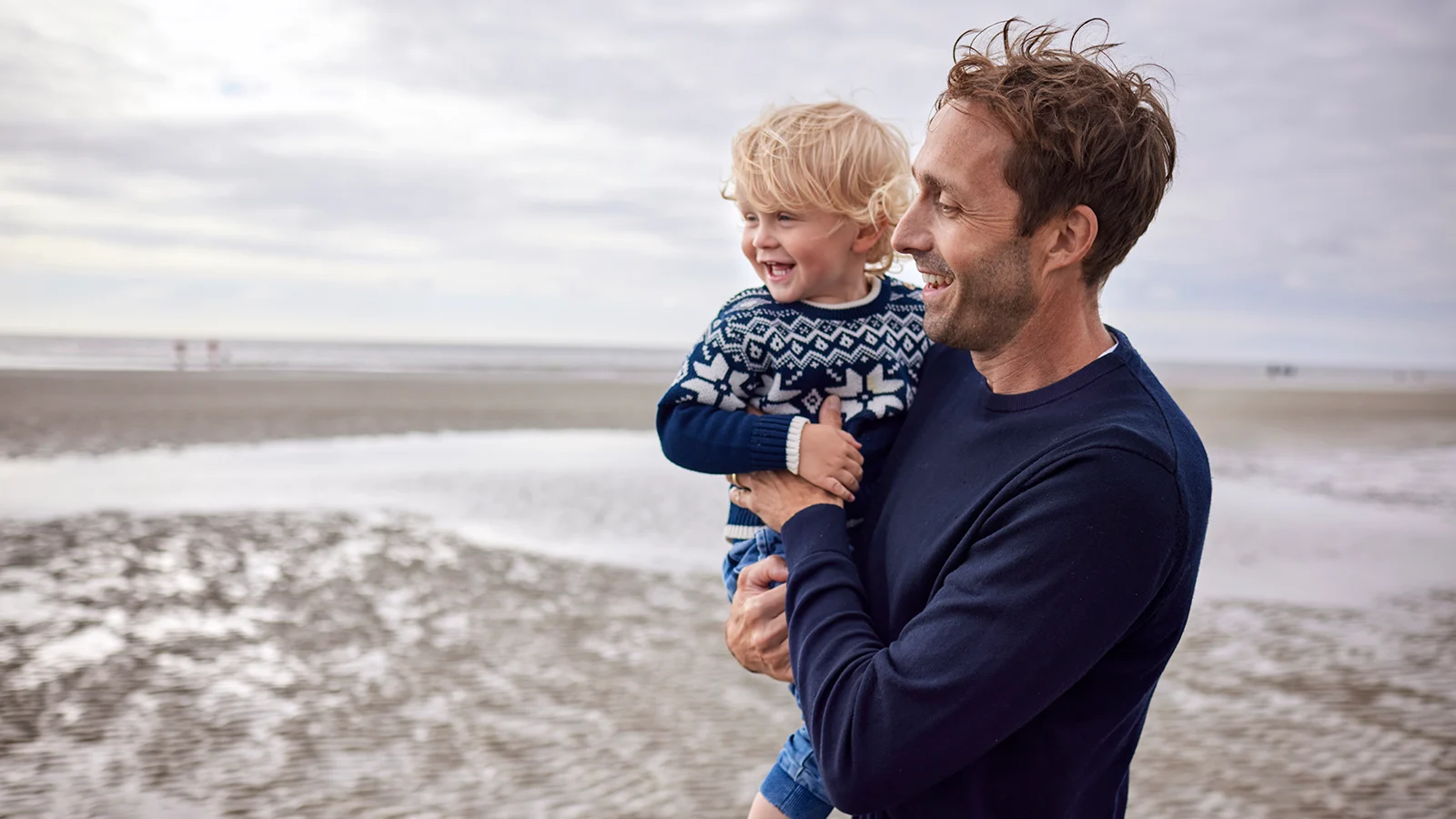 Adult carrying a young child while walking along a beach.