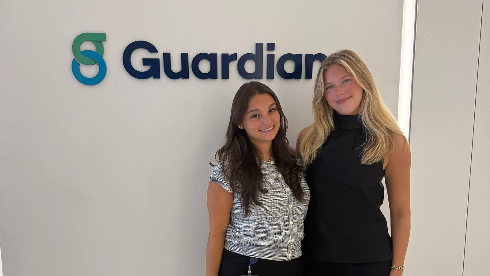 Two people pose together in front of a large Guardian logo on an office wall.