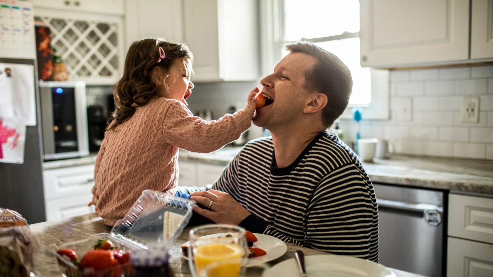 Toddler feeding her dad a strawberry in the kitchen.