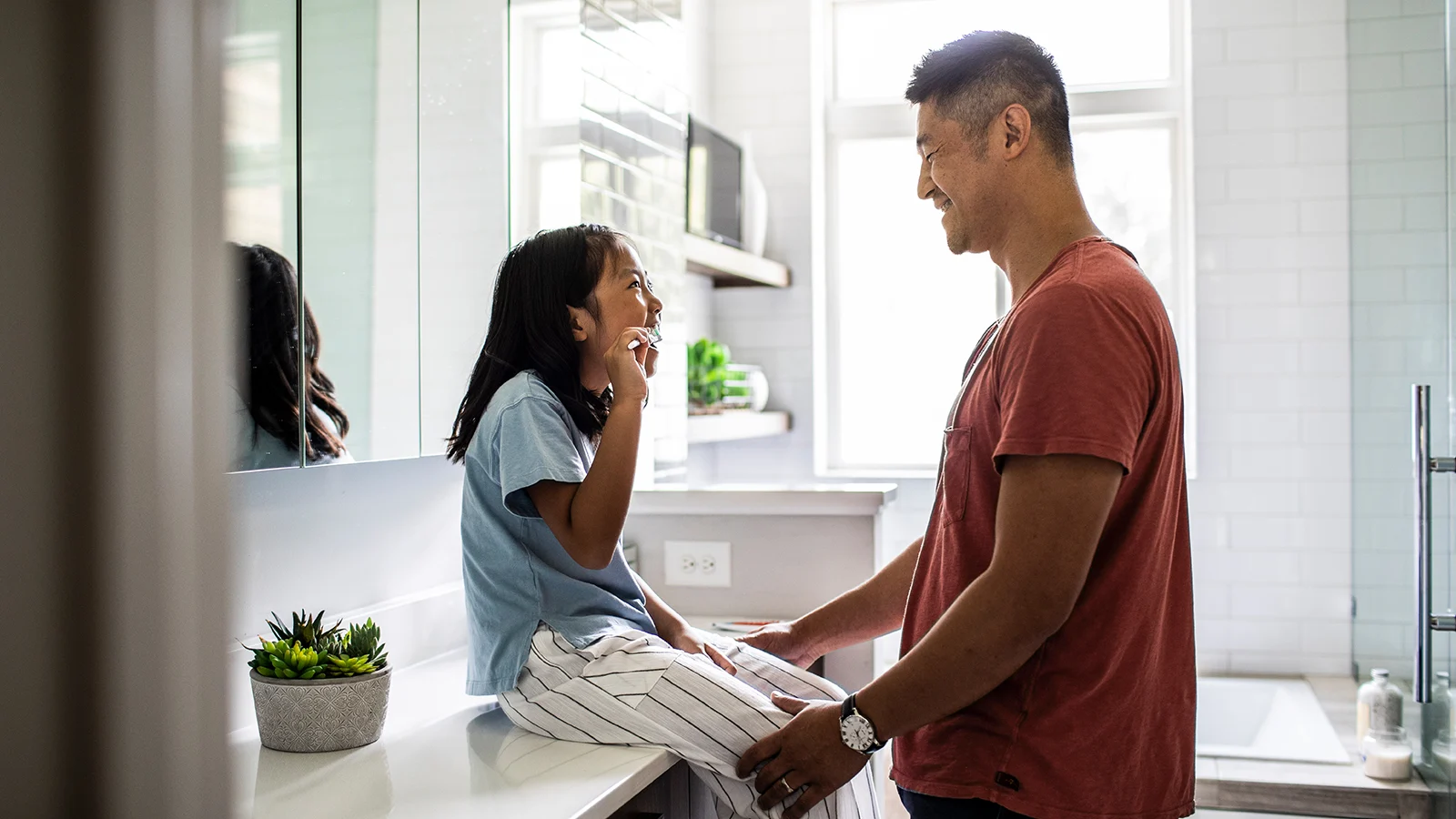 Mental wellness + oral health - young daughter brushing teeth looking at dad