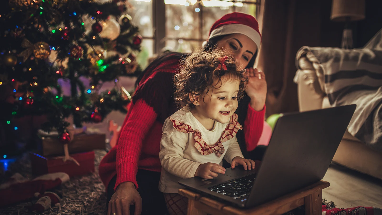 Child using a laptop with an adult wearing a Santa hat beside them near a decorated Christmas tree in a cozy holiday-themed living room.