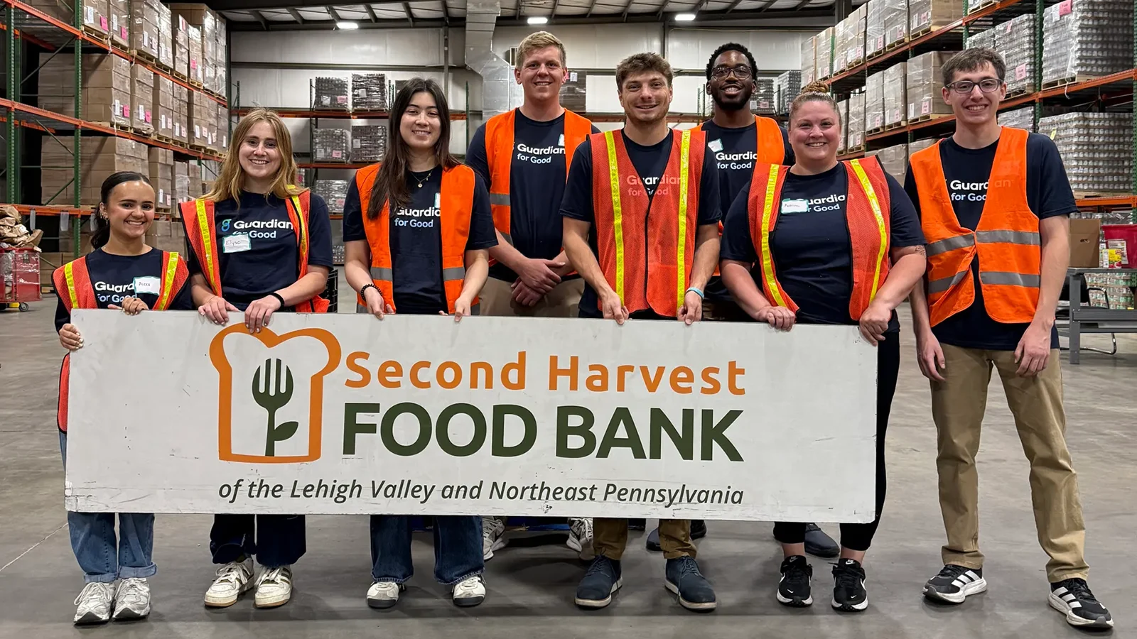 Volunteers wearing Guardian for Good shirts and safety vests pose behind a Second Harvest Food Bank sign in a warehouse.