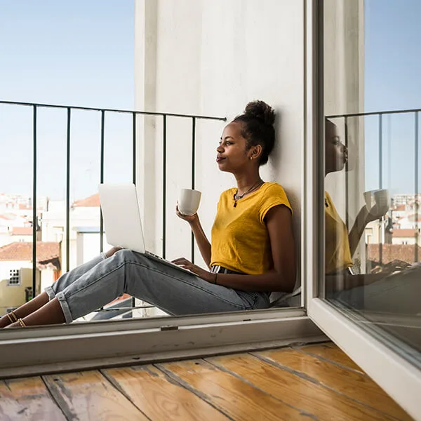 Woman sitting on her balcony with her laptop drinking from a cup