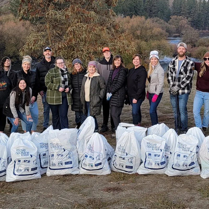 Corporate impact - A large group of Guardian colleagues stand in a line beside a river, showing off a row of trash bags they filled with trash from the riverside 