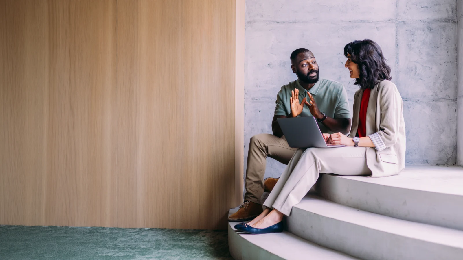 Two people seated on steps talking while using a laptop.