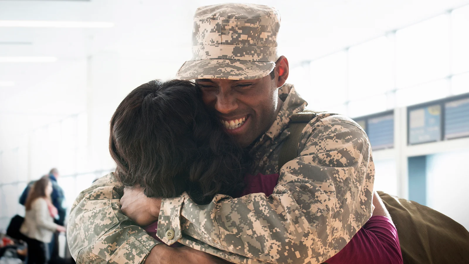 Person in military camouflage uniform hugging another person in an airport terminal.