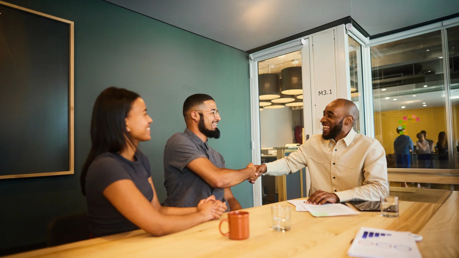 Two people shake hands across a conference table during a meeting in a modern office while a third person sits nearby.