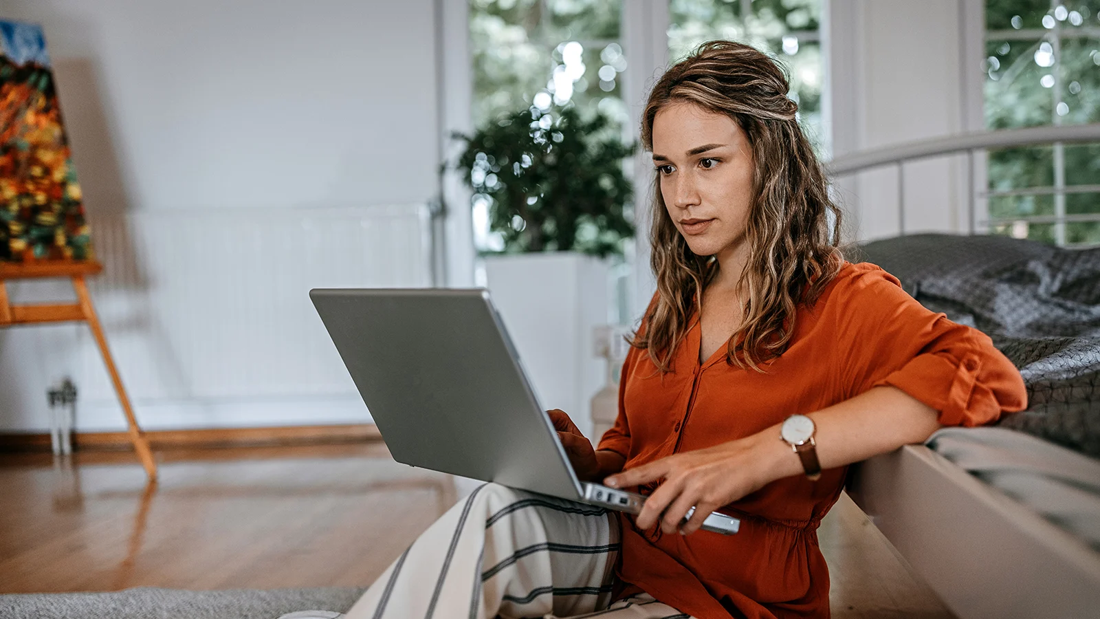 Person sitting on the floor beside a bed using a laptop in a bright room with large windows and indoor plants.