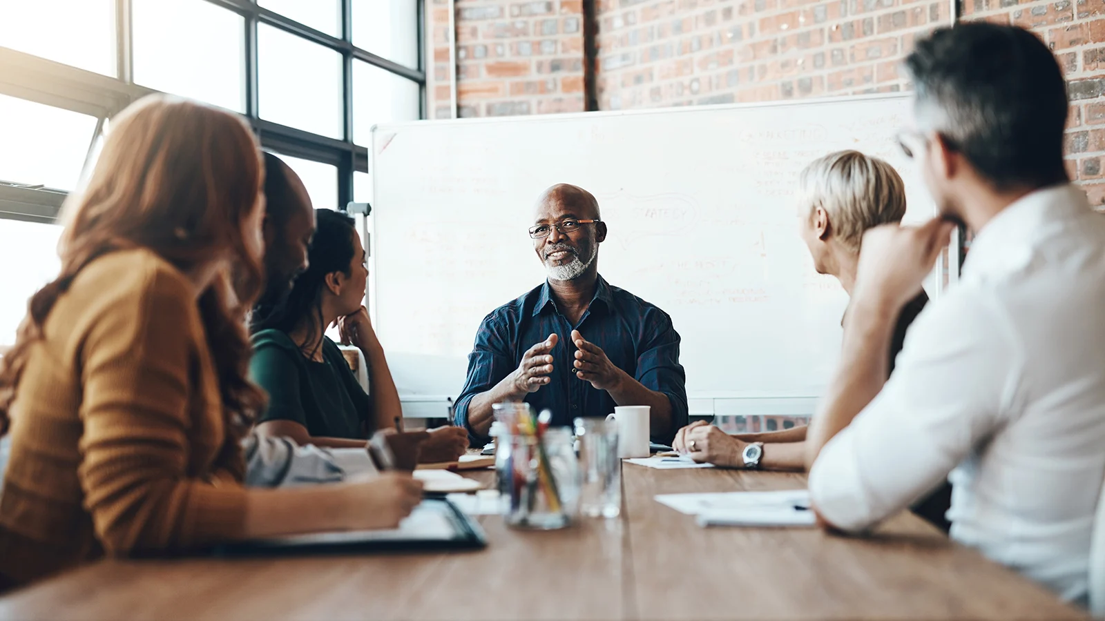 Employees seated around a conference table during a team meeting, with a presenter speaking in front of a whiteboard in a modern office.