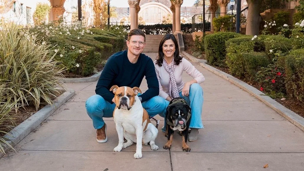 A husband and wife kneel in a landscaped garden with their two dogs.