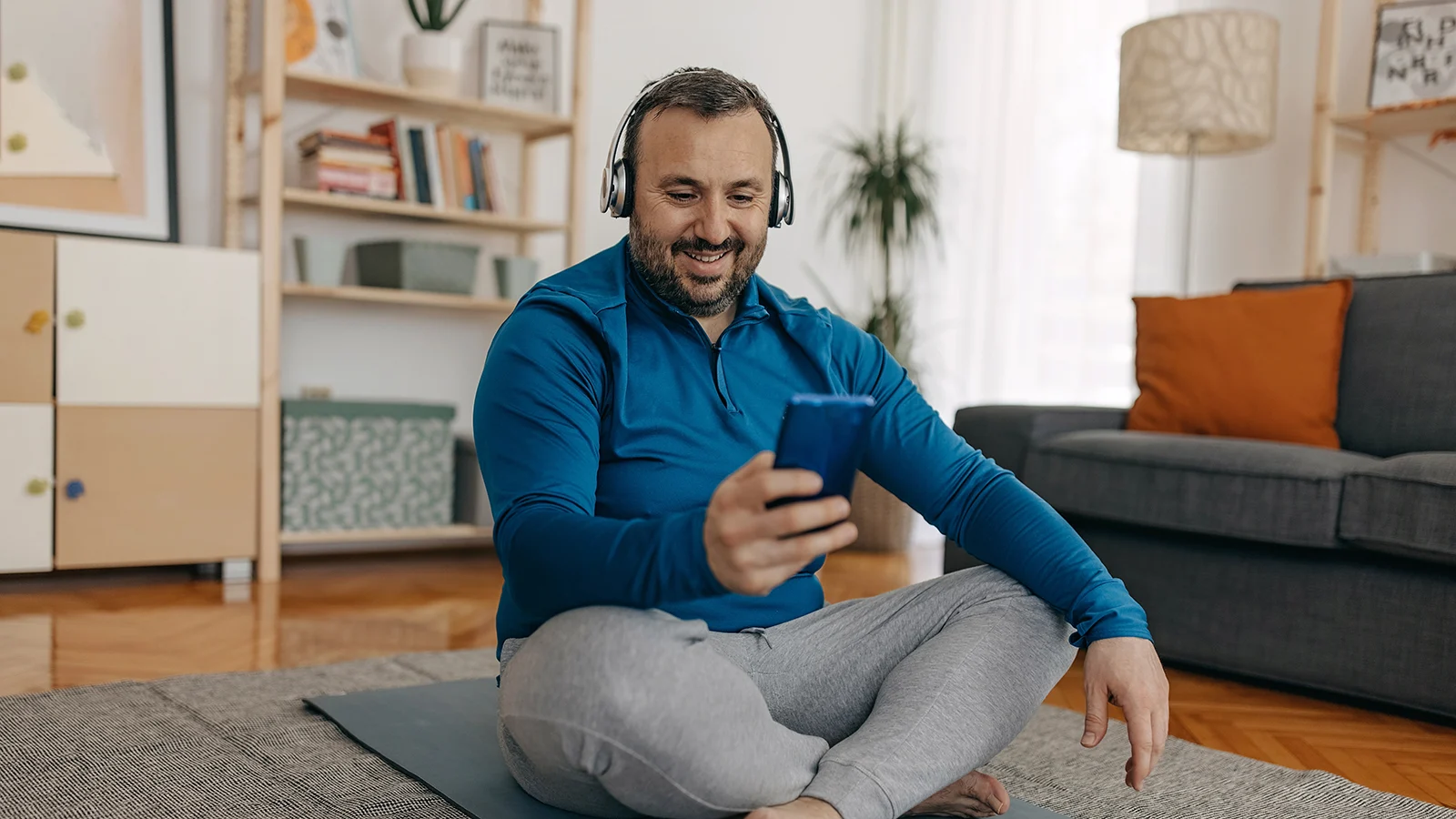 Person sitting on a yoga mat at home, wearing headphones and looking at a smartphone during a virtual workout session.