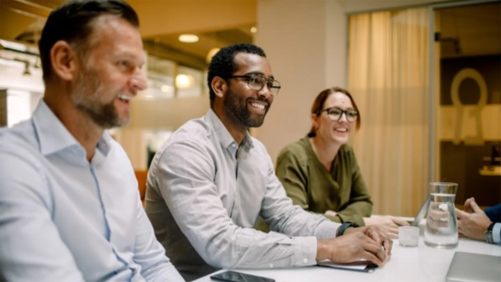 Three professionals seated at a conference table participating in a workplace discussion, with a laptop and water carafe on the table.