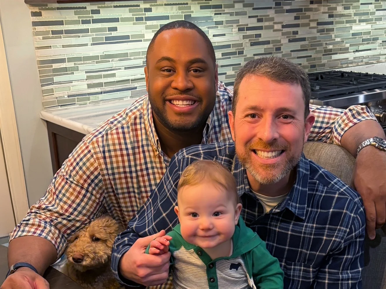 Two men smile at the camera with their baby boy and dog.