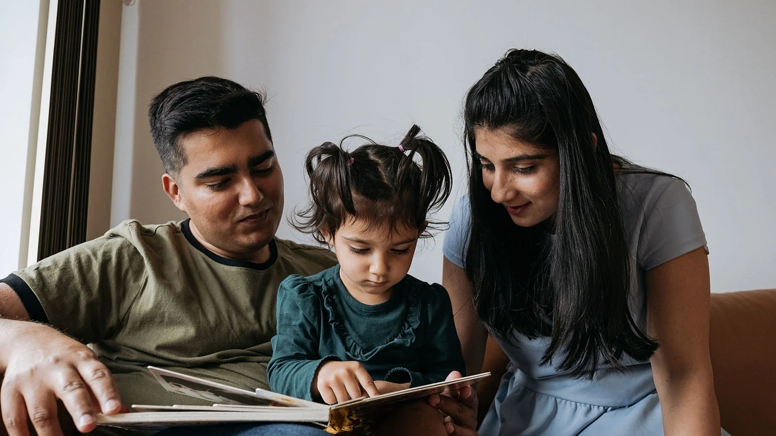 Child sitting between two adults on a couch, pointing to a page in an open book while all three read together at home.