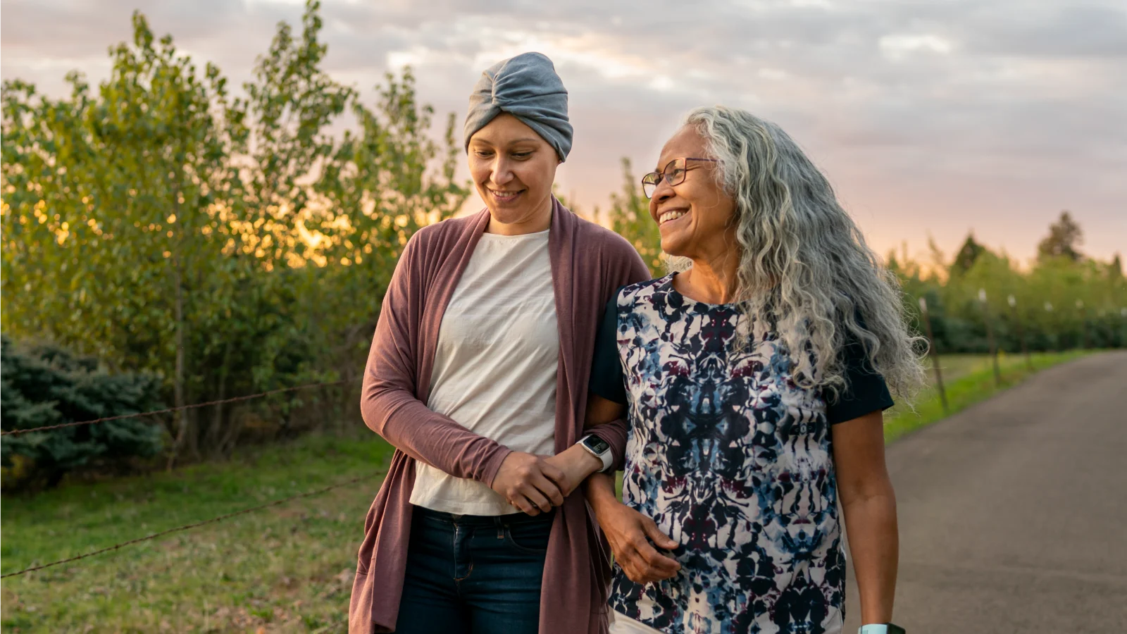 Standing Up and Stepping In: A modern look at caregivers in the US - Two women, one who's undergoing cancer treatment, walking together with linked arms. 