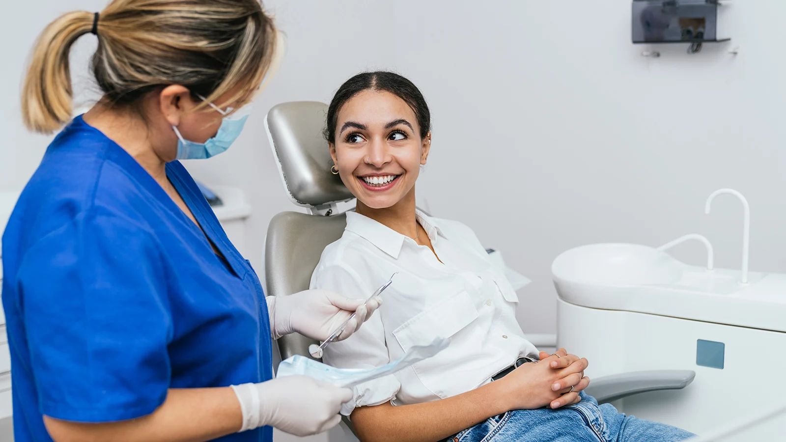 Healthcare professional in blue scrubs and protective mask speaks with a patient seated in a dental examination chair while holding dental instruments.