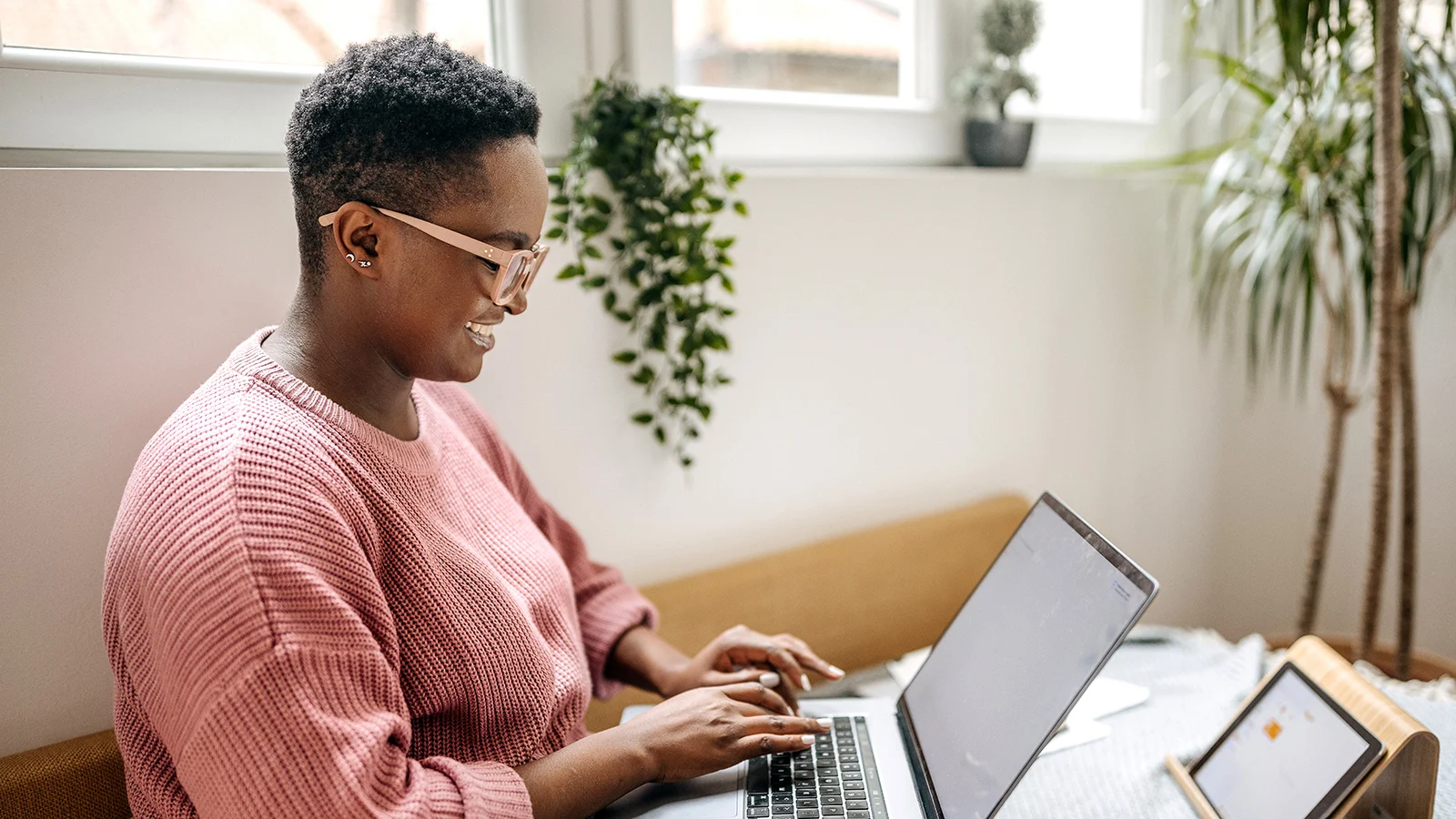 Person working on a laptop in a home office setting with natural light and indoor plants.