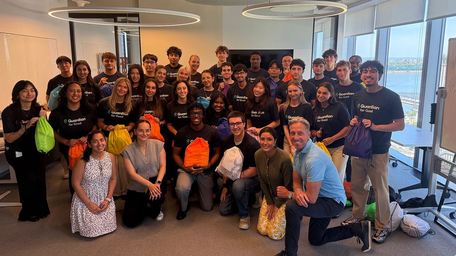 A large group poses together in an office, many wearing matching Guardian for Good shirts and holding colorful drawstring bags.