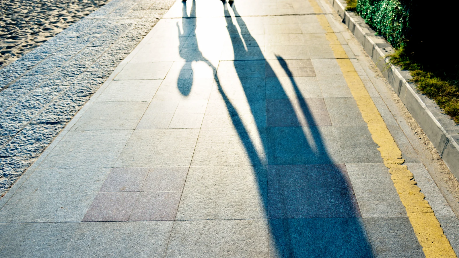 Next-Gen Wealth - Overview - Image 2 Long shadows of two people walking along a sunlit paved path beside a sandy area.