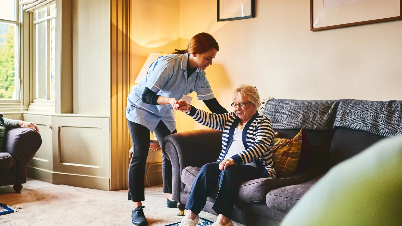 Professional caregiver helping an older woman stand up from a couch.