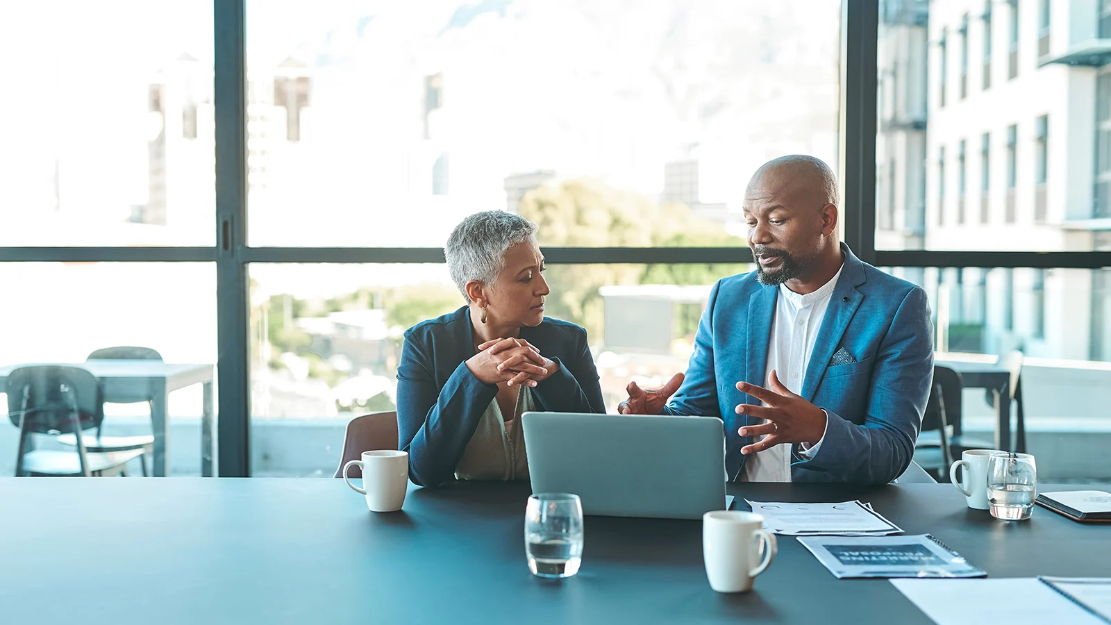 Two people meeting at a conference table with a laptop, documents, coffee mugs, and glasses of water in a sunlit office.
