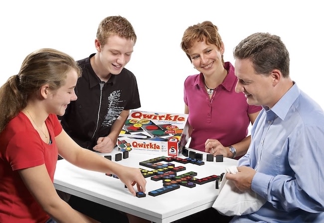 Zwei Erwachsene und zwei Kinder sitzen um einen Tisch und spielen das Spiel „Qwirkle“