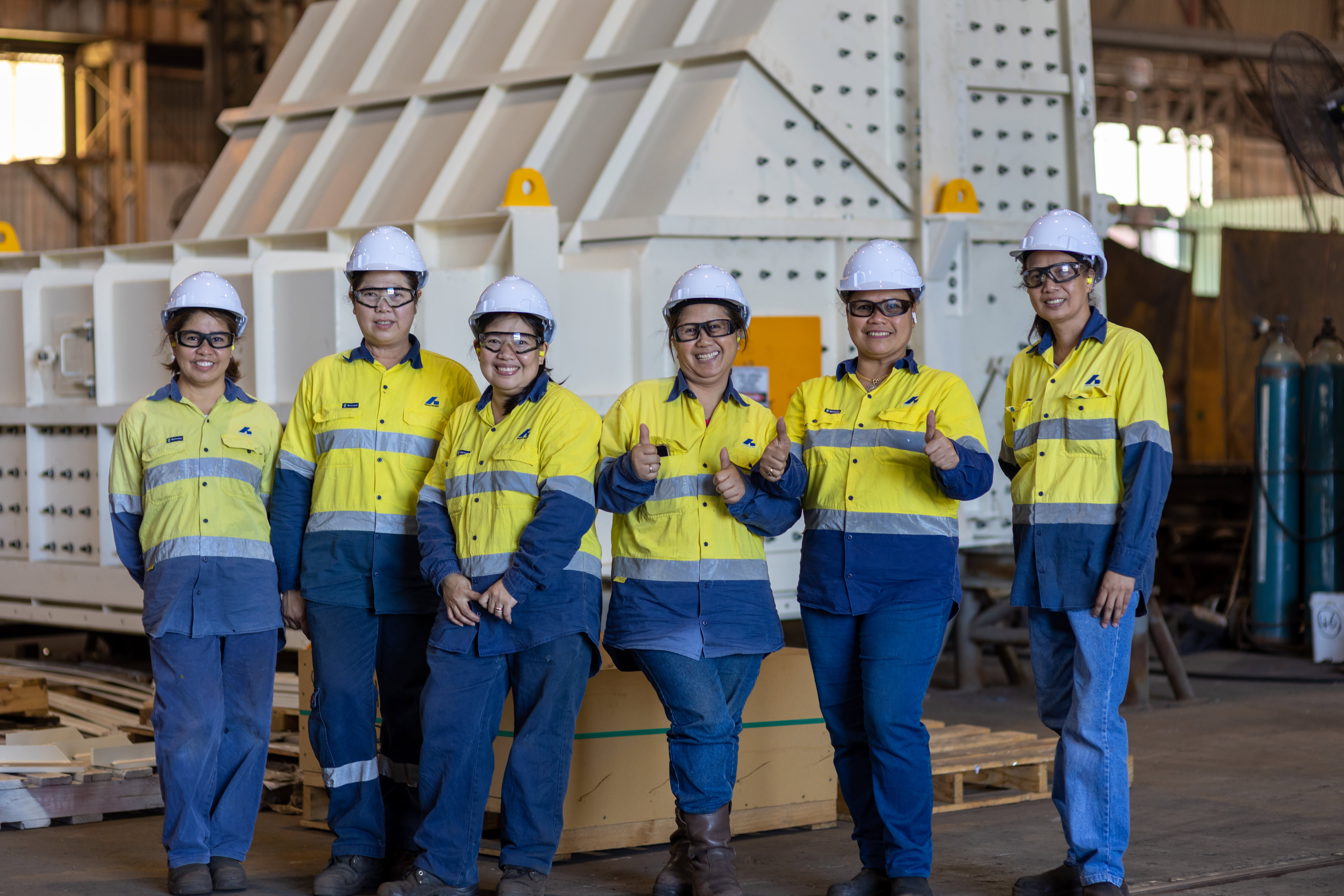 A group of six women in hi-visibility clothing stand in front of a large white mining chute component in a workshop.