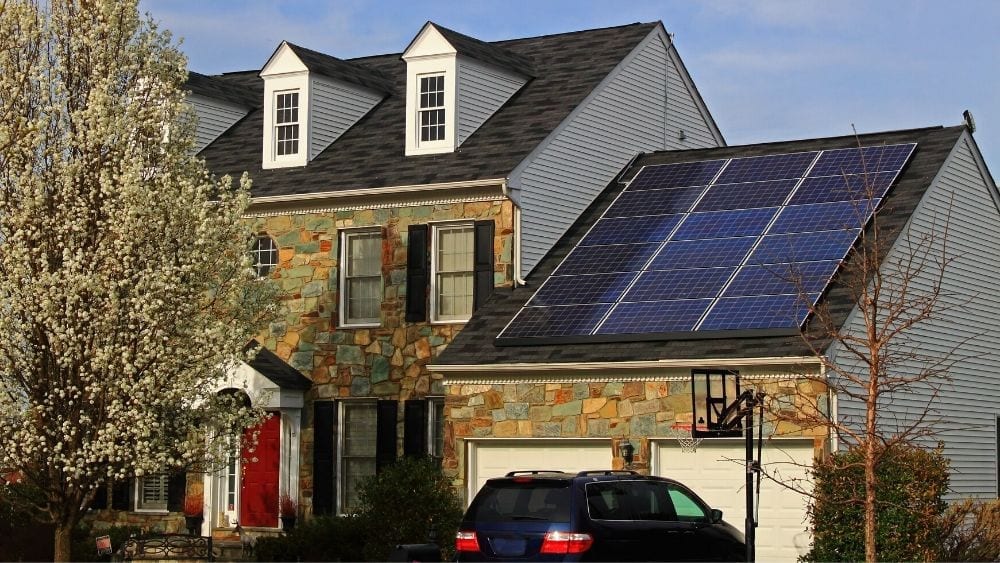 House with black roof dormer windows and solar panels above a two-car garage, with a basketball net in front
