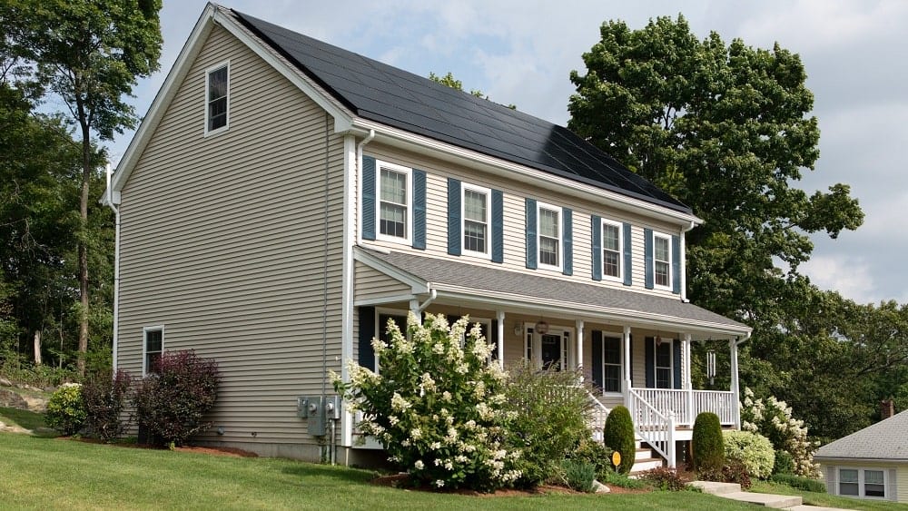 Two-story house with roof covered in solar panels, with covered front porch, and trees and landscaping