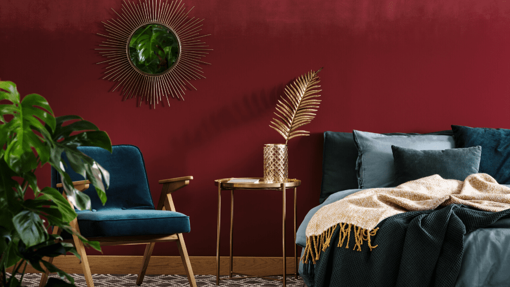 a blue chair, a gold table, and a bed with blue linens in front of a maroon wall