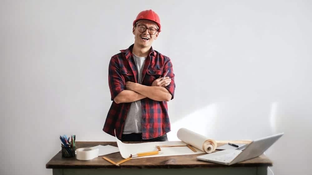 Smiling builder behind desk with papers, pencils, and a laptop.