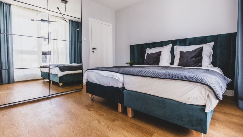 A bed against one wall with blue and white sheets. A wall of mirrors is to one side, and the floor is light brown vinyl.
