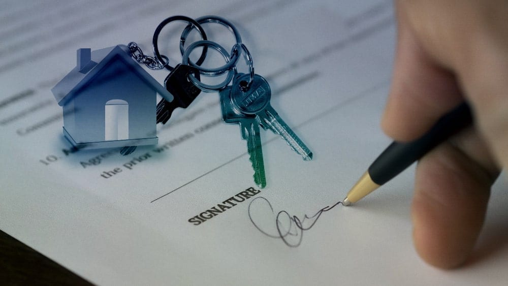 Image of a hand holding a pen and signing a name on a form with a set of keys on a house key chain to indicate selling a home