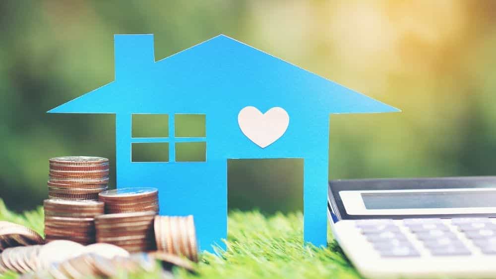 Cutout of a blue model house on a table next to a stack of coins and calculator