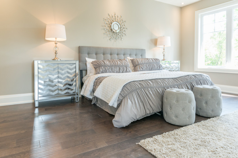 Guest bedroom with grey bedding, two grey ottomans at the foot of the bed, grey tufted headboard, large window and metallic nightstands with white lamps.