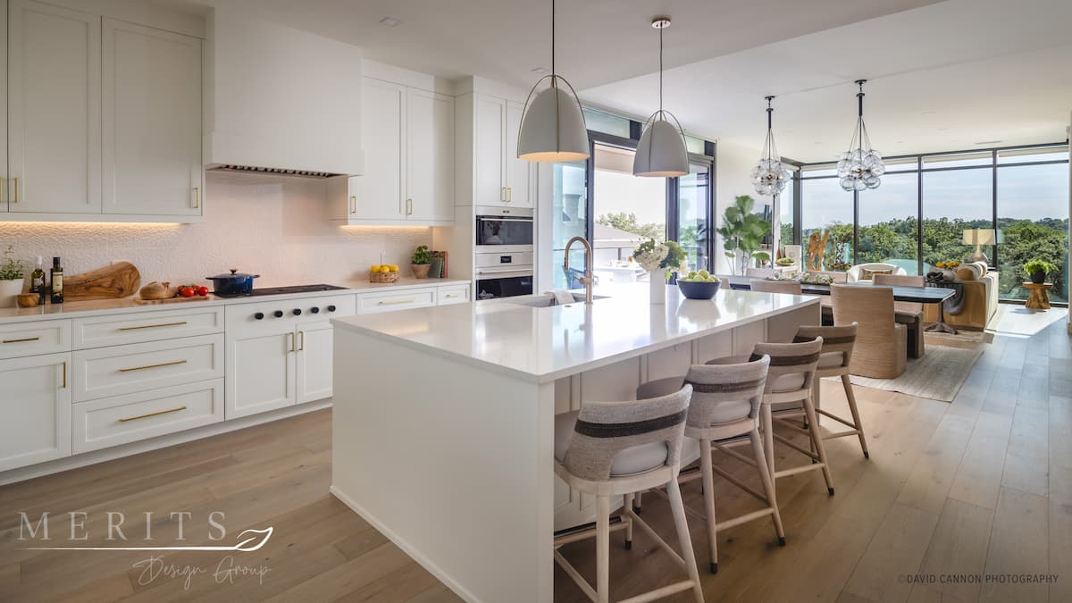 Open plan kitchen designed for wellness, with large, white kitchen island, dining table, white perimeter cabinetry and floor-to-ceiling windows.