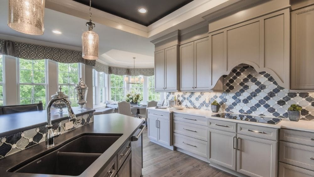 Modern kitchen with a grey, black, and white color scheme. A patterned tile backsplash is behind the stove, there is a kitchen island with a double sink, and windows line the wall by the breakfast table.