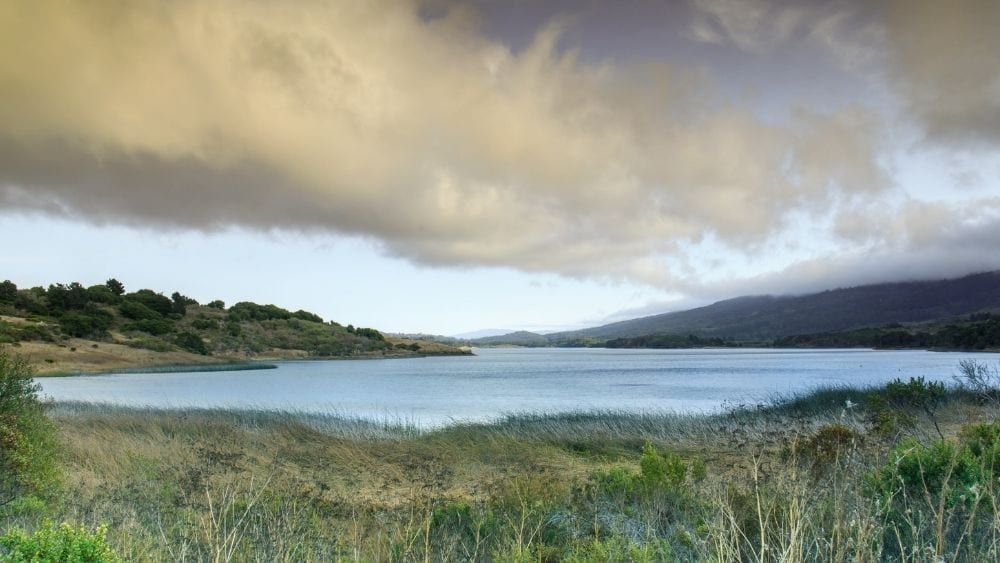 An open field with a lake in the middle and mountains rising on either side. The clouds above are overcast.