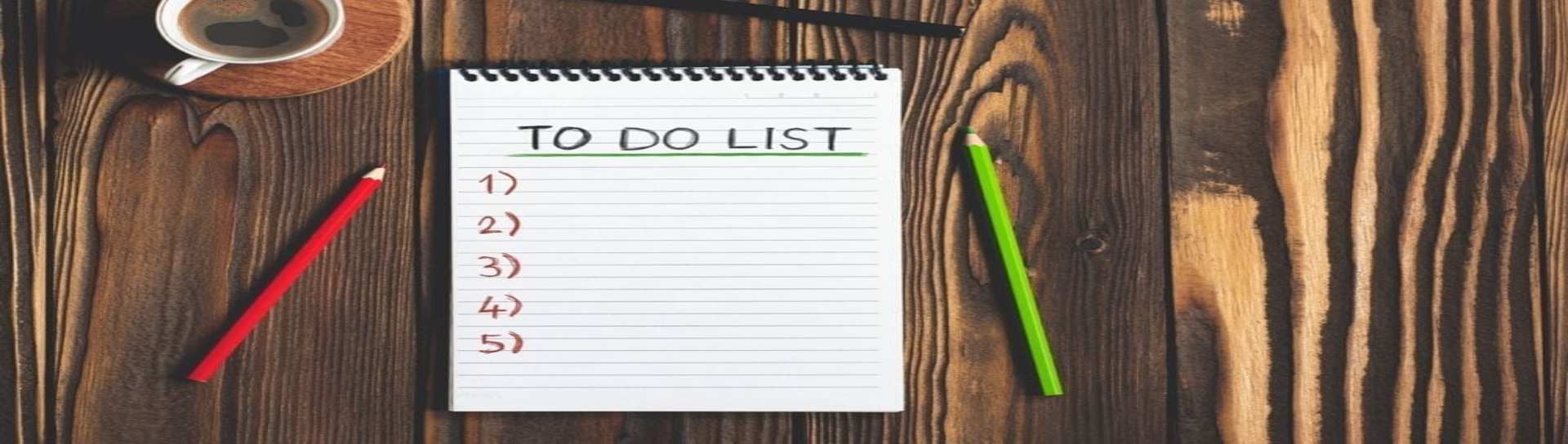 A pad of white paper open to a to-do list, with numbered points, on a wood table next to a green pencil, red pencil and coffee cup