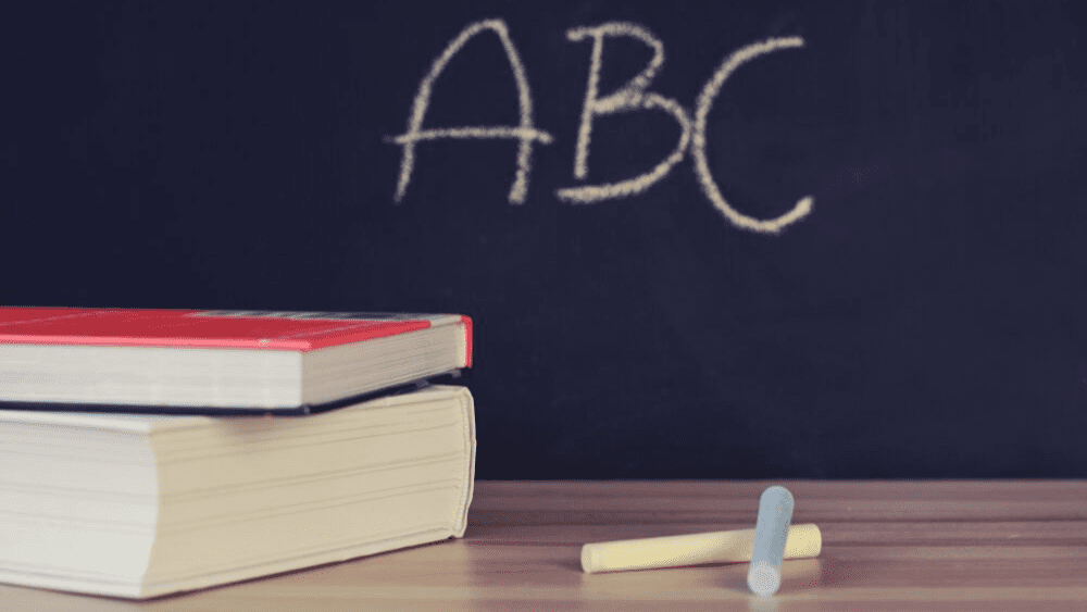Two books sit next to a pile of chalk in front of a blackboard reading “ABC”