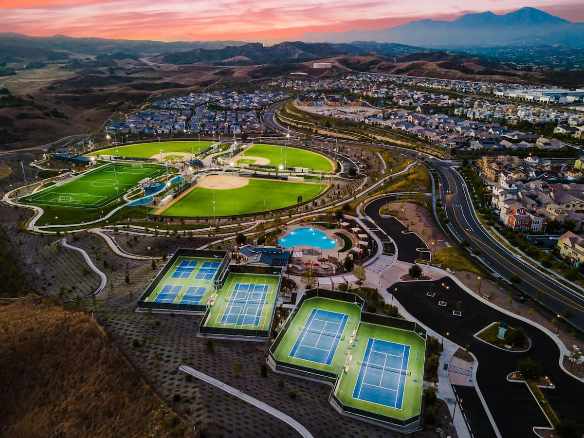 Multiple tennis courts in the foreground with green space, hills, and a sunset in the background