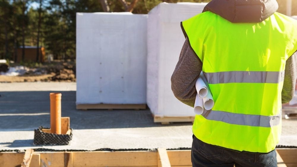 An architect in a construction vest looking at a foundation that’s been laid for a house.