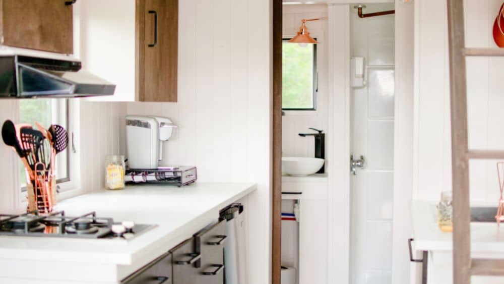 Kitchen in a tiny home with a small white counter and bathroom behind, with a brown ladder against the wall.