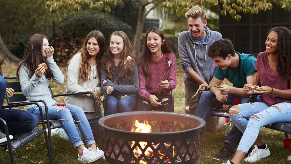 7 people sitting around the fire pit, laughing and roasting marshmallows