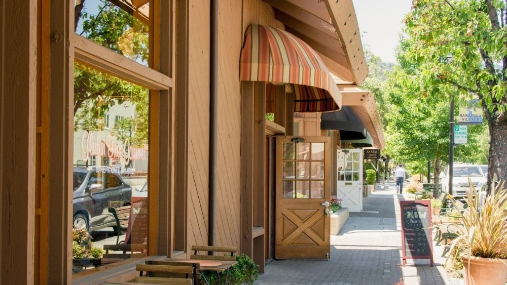 Red clay buildings with awnings and large window fronts line a street. Patio chairs and tables are in front of some of the buildings.