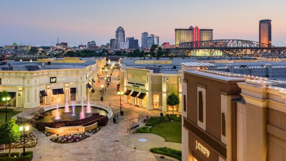 A city square with a fountain and shops surrounding it; the city skyline is in the background.