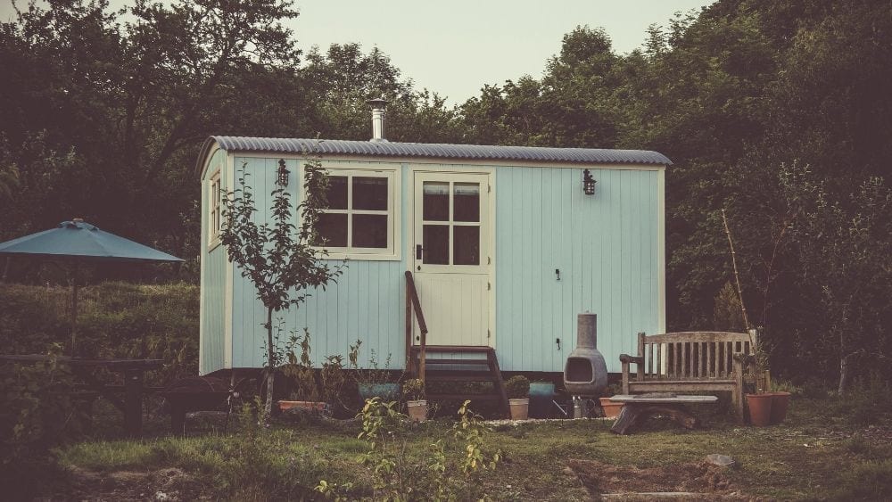 Small blue house with a white door sits behind a wooden bench and a picnic table with a dark blue umbrella.