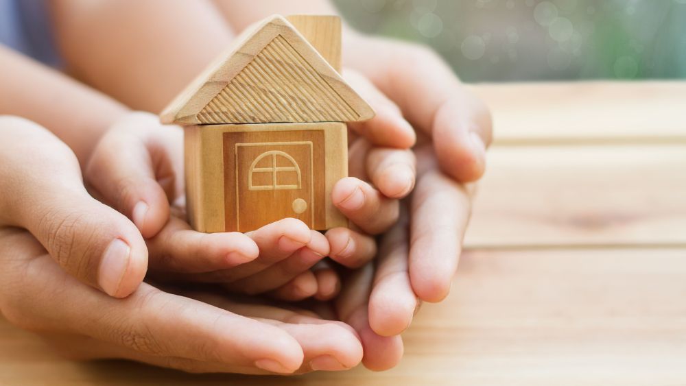 Parents hands cradling small child's hands holding a wooden model of a house